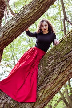 beautiful woman in a red dress sitting on a trunk in the spring forest