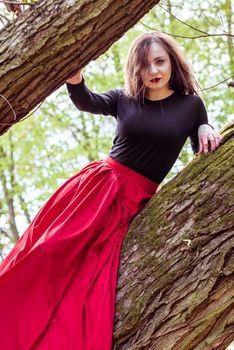 beautiful woman in a red dress sitting on a trunk in the spring forest