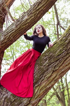 beautiful woman in a red dress sitting on a trunk in the spring forest