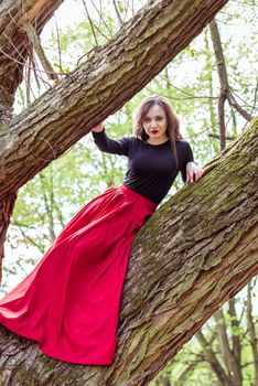 beautiful woman in a red dress sitting on a trunk in the spring forest