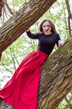 beautiful woman in a red dress sitting on a trunk in the spring forest