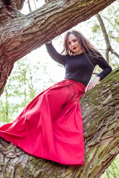 beautiful woman in a red dress sitting on a trunk in the spring forest