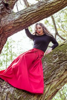 beautiful woman in a red dress sitting on a trunk in the spring forest