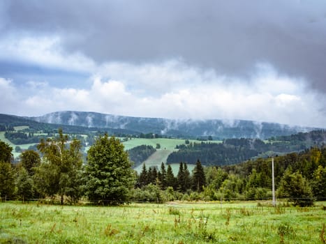 Dramatic cloudy misty landscape. mountain meadow and forests, both deciduous broadleaved and needle coniferous trees, electric overhead power line and horizon covered in clouds and mist, Czech republic, central Europe, Orlicke hory