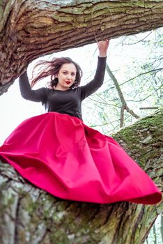 beautiful young woman in a red skirt sitting on a tree trunk in spring forest