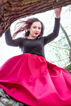 beautiful young woman in a red skirt sitting on a tree trunk in spring forest