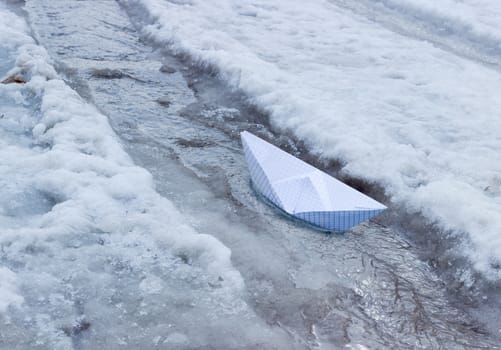 Toy paper boat made of squared paper in the stream of melt water among ice and wet snow
