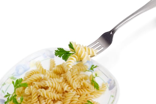 Fragment of the stainless steel fork with several cooked spiral pasta and the parsley twig over of a dish with the same pasta on a light background
