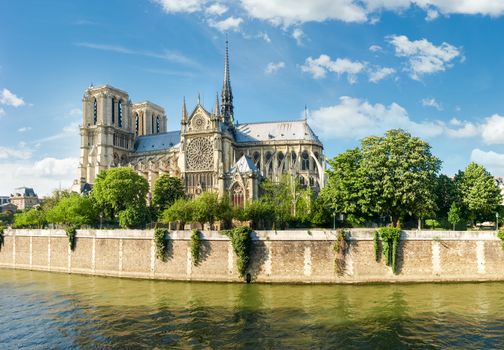 View of the southern facade of the Cathedrale Notre-Dame de Paris from the Seine with the embankment of the river in the foreground against the sky with clouds
