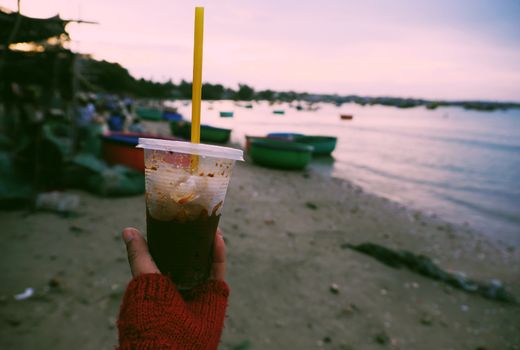 Woman hand hold coffee cup up to sky at beach in morning, quick cafe time on the way in summer journey to enjoy life