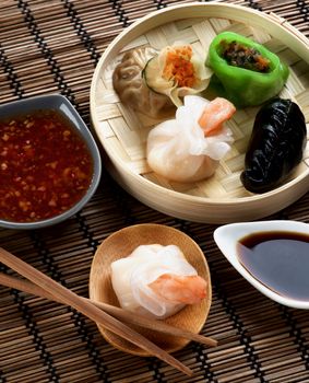 Assorted Dim Sum in Bamboo Steamed Bowl and Tori with Shrimp on Wooden Plate with Red Chili and Soy Sauces and Chopsticks closeup on Straw Mat background