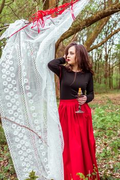 mystical witch woman in a red skirt with a candle hidden behind the curtains in spring forest