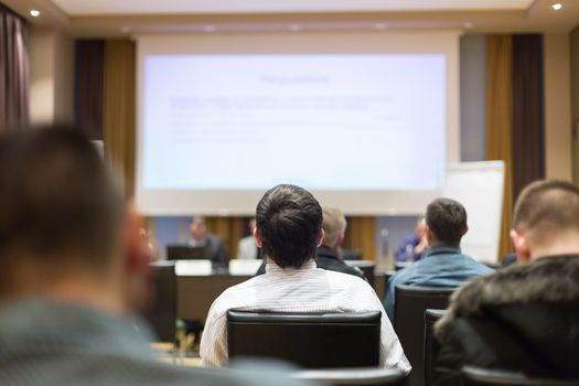 Business and entrepreneurship symposium. Speaker giving a talk at business meeting. Audience in the conference hall. Rear view of unrecognized participant in audience.