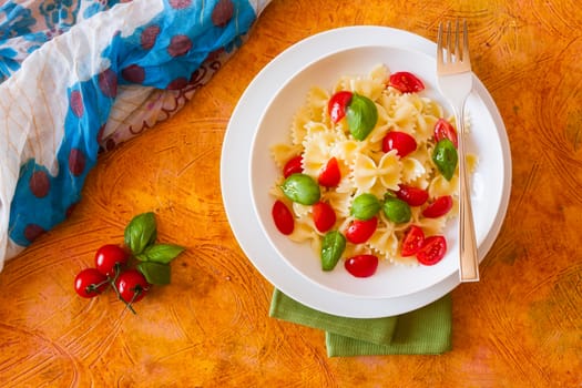Farfalle pasta with cherry tomatoes and basil seen from above over a colored background