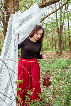 mystical witch woman in a red skirt with a lantern hidden behind the curtains in spring forest