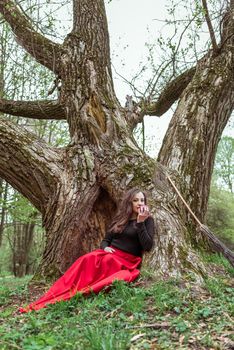 mystical witch woman in red dress sitting under a old tree in the spring forest