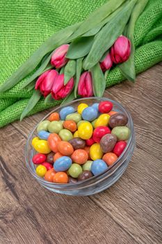 Colorful candies in a glass bowl with red tulips on a wooden background