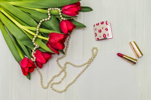 red tulips in the necklace, lipstick and a white sheet of paper on the background of white wooden board