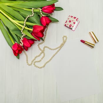red tulips in the necklace, lipstick and a white sheet of paper on the background of white wooden board