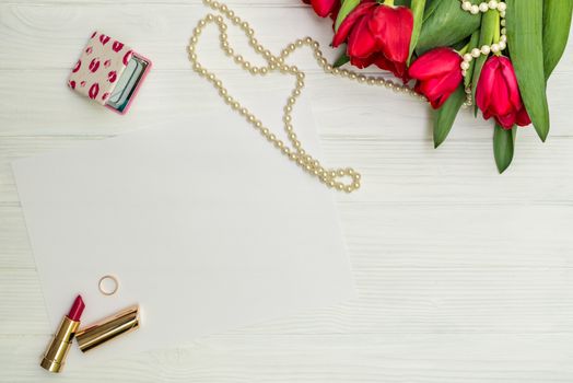 red tulips in the necklace, lipstick, one ring and a white sheet of paper on the background of white wooden board