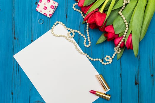 red tulips in the necklace, lipstick, one ring and a white sheet of paper on the background of blue wooden board