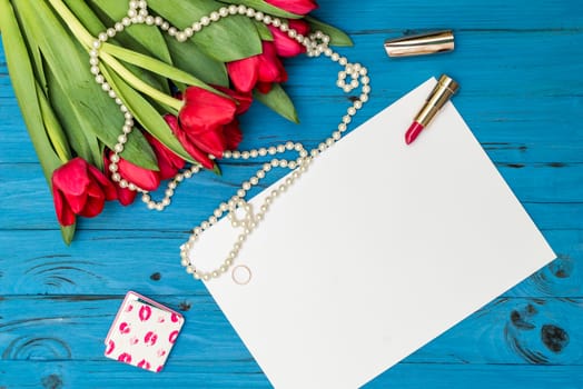 red tulips in the necklace, lipstick, one ring and a white sheet of paper on the background of blue wooden board