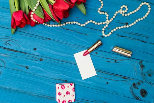 red tulips in the necklace, lipstick and a white card on the background of blue wooden board