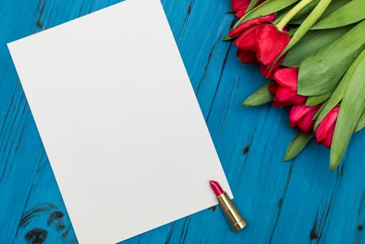 red tulips, lipstick and a white sheet of paper for your greetings on the background of blue wooden board