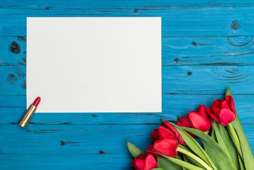 red tulips, lipstick and a white sheet of paper for your greetings on the background of blue wooden board