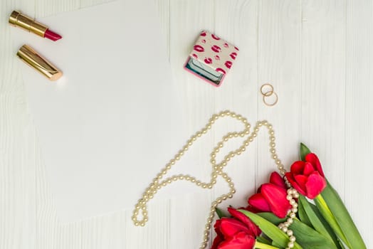 red tulips in the necklace, lipstick, two rings and a white sheet of paper on the background of white wooden board