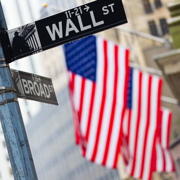 Wall street sign in New York with American flags and New York Stock Exchange in background.