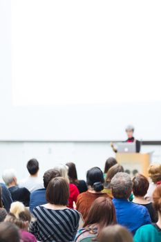 Business and entrepreneurship symposium. Female speaker giving a talk at business meeting. Audience in conference hall. Rear view of unrecognized participant in audience. Copy space on whitescreen.