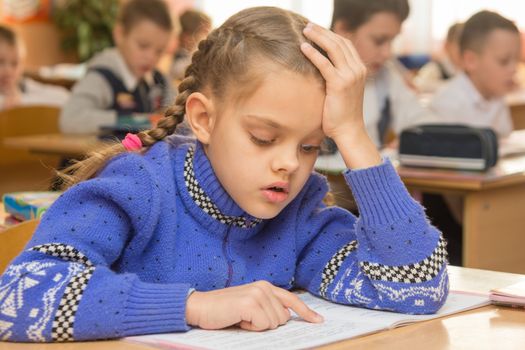 First-grader at a lesson reads the text line by line leading a finger, in the background other students