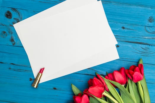 red tulips, lipstick and a white sheet of paper for your greetings on the background of blue wooden board