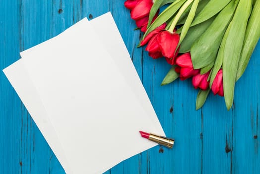 red tulips, lipstick and a white sheet of paper for your greetings on the background of blue wooden board