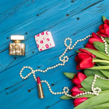 red tulips in the necklace, lipstick and bottle of perfume on the background of blue wooden board