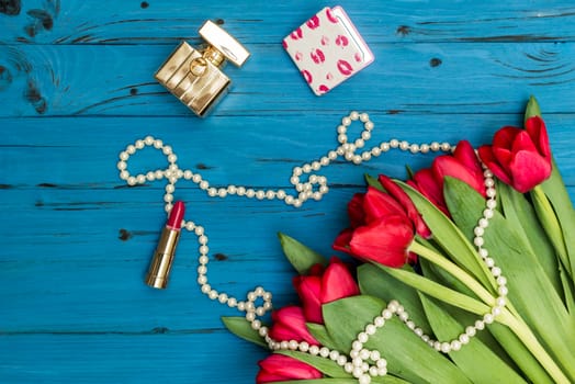 red tulips in the necklace, lipstick and bottle of perfume on the background of blue wooden board