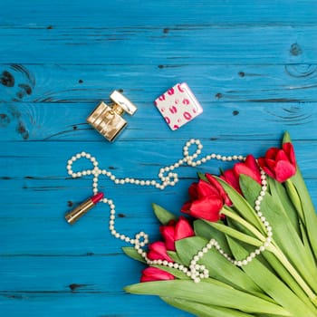 red tulips in the necklace, lipstick and bottle of perfume on the background of blue wooden board