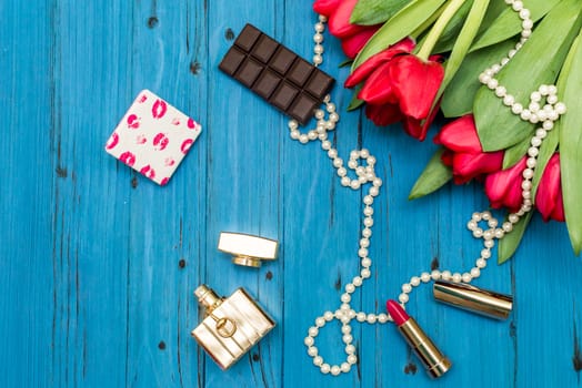 red tulips in the necklace, lipstick, bottle of perfume and piece of chocolate on the background of blue wooden board