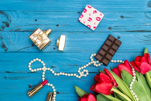 red tulips in the necklace, lipstick, bottle of perfume and piece of chocolate on the background of blue wooden board