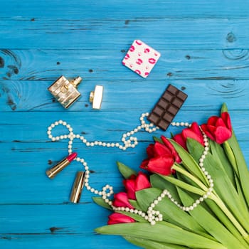 red tulips in the necklace, lipstick, bottle of perfume and piece of chocolate on the background of blue wooden board