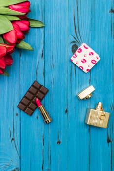 red tulips in the necklace, lipstick, bottle of perfume and piece of chocolate on the background of blue wooden board