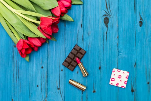 red tulips in the necklace, lipstick and piece of chocolate on the background of blue wooden board