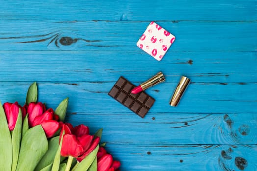 red tulips in the necklace, lipstick and piece of chocolate on the background of blue wooden board