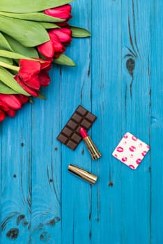 red tulips in the necklace, lipstick and piece of chocolate on the background of blue wooden board