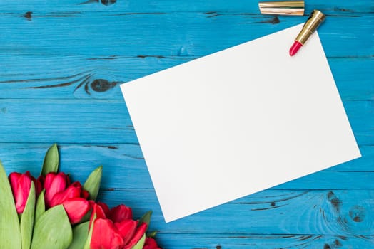 red tulips, lipstick and a white sheet of paper for your greetings on the background of blue wooden board