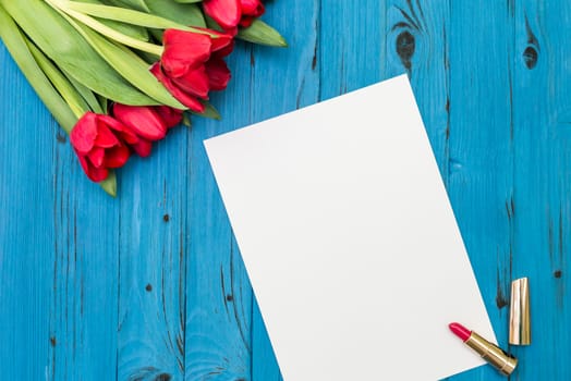 red tulips, lipstick and a white sheet of paper for your greetings on the background of blue wooden board