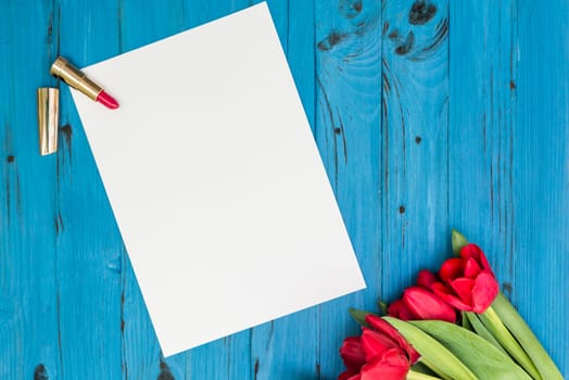 red tulips, lipstick and a white sheet of paper for your greetings on the background of blue wooden board