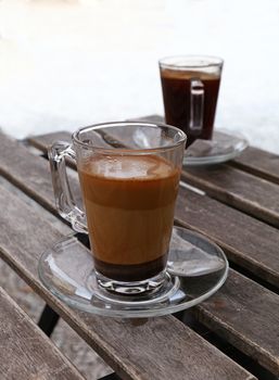 Two full Turkish freshly brewed natural ground coffee, black and with milk, in transparent full glass cups with saucer and metal spoons over vintage wooden table, close up, high angle view