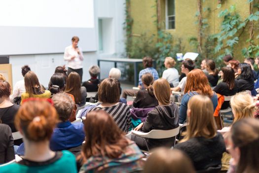 Female speaker giving presentation in lecture hall at university workshop. Audience in the conference hall. Rear view of unrecognized participant in audience. Scientific conference event.